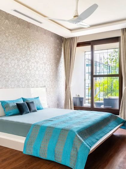 This bedroom combines a patterned accent wall with clean, modern furniture. The large window allows for ample natural light, which highlights the rich wooden flooring and the vibrant blue tones of the bedding, creating a balanced and restful atmosphere.