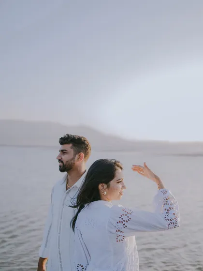 A beautiful, candid moment of the couple by the sea at sunset. The soft light and their relaxed posture create a serene and romantic atmosphere.