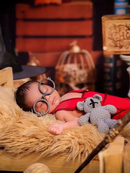 This little wizard is fast asleep, holding his wand and cuddling a teddy bear. The setup includes vintage props like a globe and birdcage to enhance the magical theme.