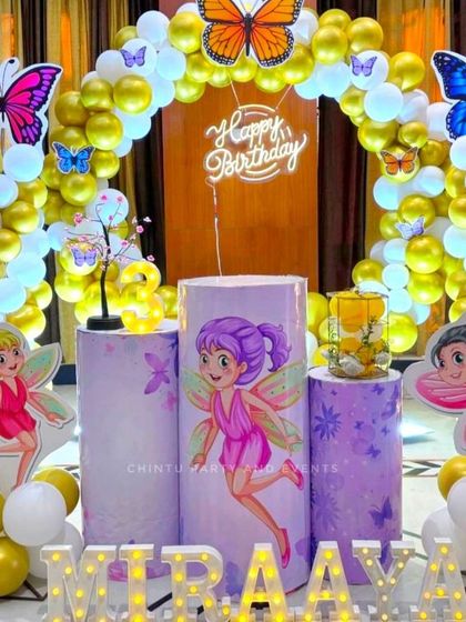 A butterfly fairy theme for a girl's birthday, combining fairy character cutouts with butterfly accents. The setup includes a balloon arch, themed pedestals, and light-up marquee letters spelling the name 'Miraaya'.