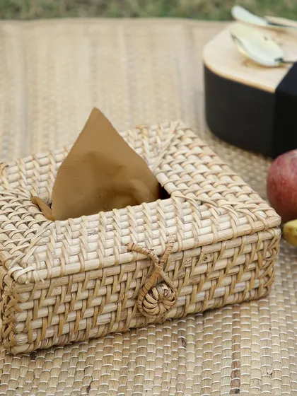 A rectangular tissue box cover, intricately woven from rattan, shown in a picnic setting. This stylish accessory elevates a simple household item into a decorative piece, perfect for any room.