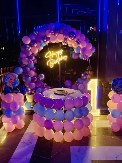 A Frozen-themed first birthday party decoration. The setup features a balloon arch in Elsa's signature colors, a character cutout, a "Happy Birthday" neon sign, and a light-up number "1".