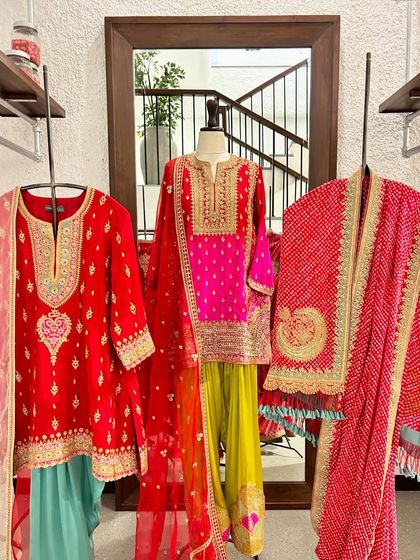 A trio of brightly colored ethnic outfits, featuring a red embroidered kurta, a pink and yellow sharara set, and a red bandhani dupatta, perfect for a Mehendi or Haldi ceremony.