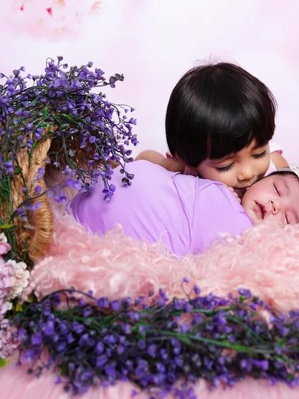 A tender moment between an older sister and her newborn sibling, who is nestled in a basket of purple and pink flowers.
