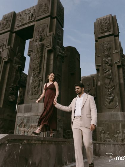 A couple explores the monumental Chronicle of Georgia, holding hands. The scale of the carved stone pillars dwarfs them, creating a powerful image of two people on a grand journey together.