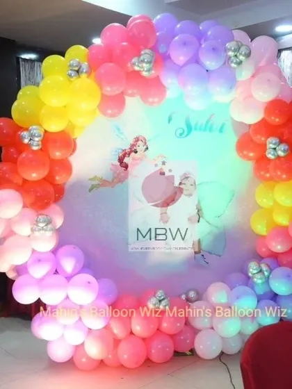 A whimsical fairy-themed birthday photo booth. The circular backdrop features fairy silhouettes and is framed by a vibrant, multi-colored balloon ring.