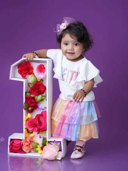 A colorful and sweet first birthday portrait. The little girl stands next to a floral number '1' in a rainbow skirt, creating a cheerful and bright image against the purple background.