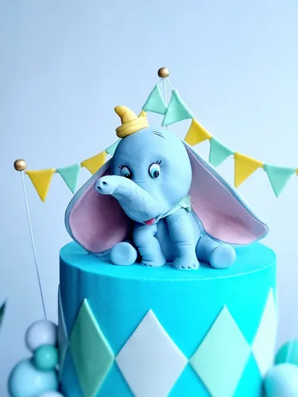 A close-up of a Dumbo-themed carnival cake. The Dumbo topper is hand-sculpted from gumpaste, and the tier features a clean, geometric argyle pattern.
