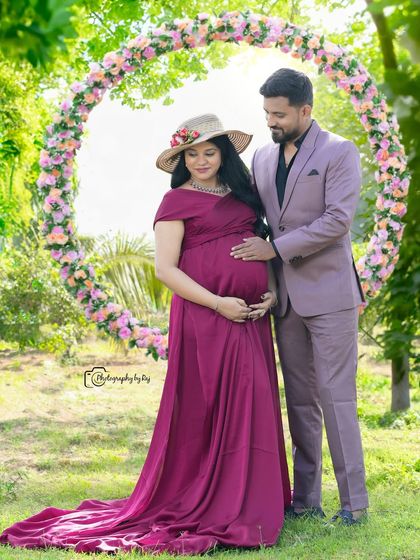 A happy couple's portrait in front of a circular floral prop. The mother-to-be is wearing a wine-colored gown and a hat, perfect for a sunny day shoot.