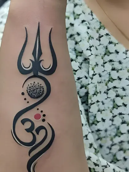 A creative forearm tattoo that combines the Trishul and Om symbols in a flowing, artistic design. The use of dotwork and a red bindi adds a unique touch.