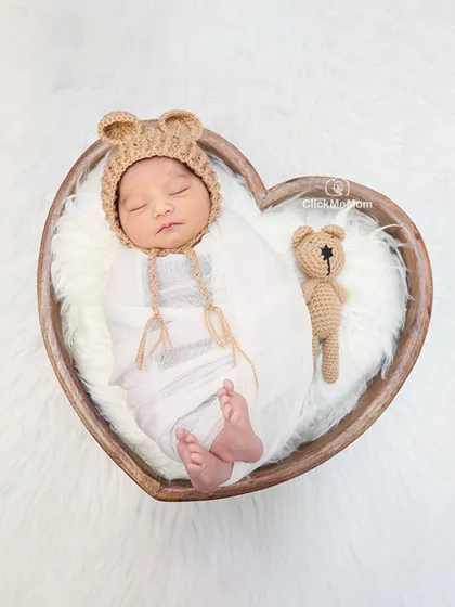 A sweet and simple portrait. This baby is wearing a teddy bear hat and is curled up inside a heart-shaped wooden bowl, creating a truly heartwarming image.