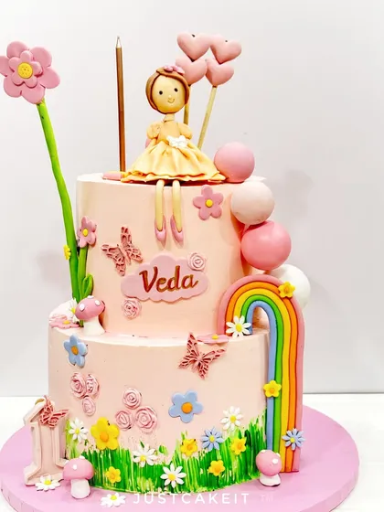 A lovely two-tier garden-themed cake for a first birthday. It features a little doll, a rainbow, and lots of hand-piped flowers and butterflies.