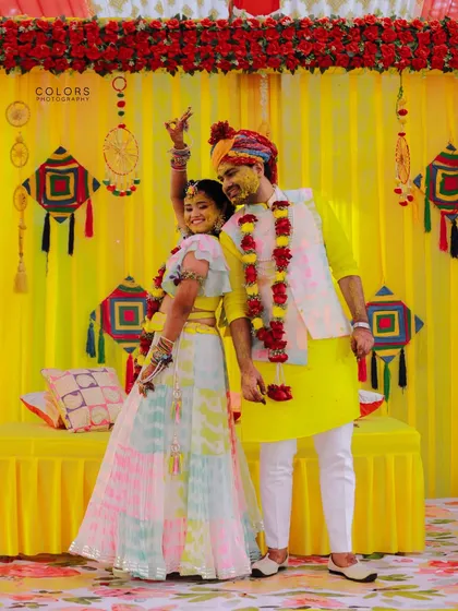 A happy couple strikes a pose during their colorful Haldi ceremony, set against a vibrant yellow backdrop with kite decorations.