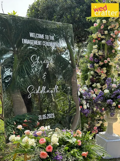 A modern mirrored welcome sign for Shreya and Siddhanth's engagement, reflecting the surrounding palm trees and flanked by lush pastel floral arrangements.