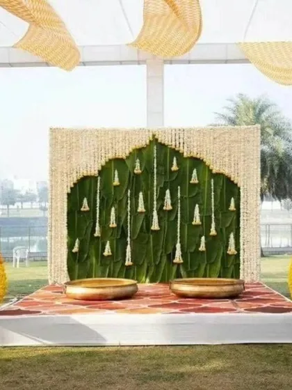 An outdoor Haldi ceremony stage featuring a traditional backdrop of banana leaves and jasmine strings, set within a carved wooden frame. The setup is completed with large brass urlis for the ceremony.
