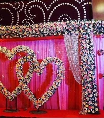 A stage decoration featuring two interlocking hearts made of flowers. This is a popular design for anniversaries and ring ceremonies, set against a rich red and pink backdrop.