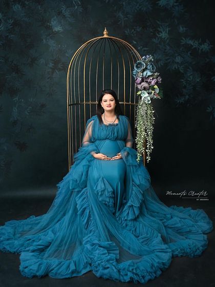 A regal portrait of the mom-to-be in a deep blue gown, seated on a chair beside the golden birdcage, creating a look of serene elegance.