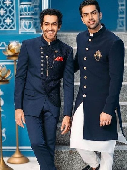 Two stylish groomsmen or brothers of the groom. One wears a navy blue asymmetrical Indo-western suit with a brocade vest, while the other is in a classic navy bandhgala. These are great coordinated looks for the wedding party.