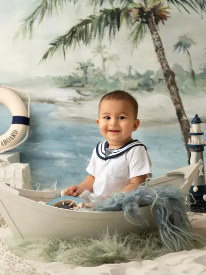 A cute little sailor smiles from his boat in this nautical-themed studio setup. The beach backdrop and props create a fun seaside adventure.