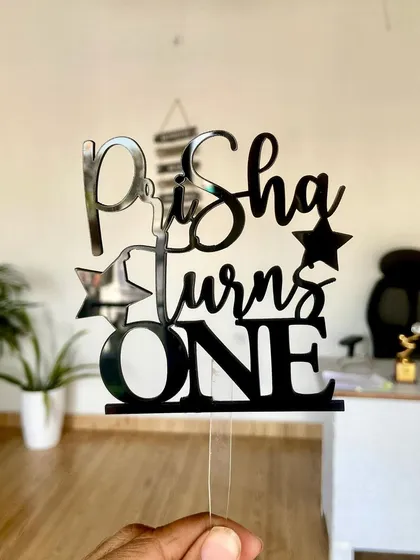 A clear shot of the "Prisha turns ONE" topper in black acrylic, showcasing its playful font and star details.