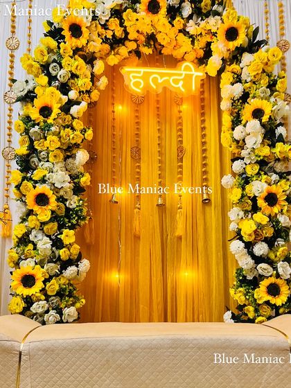 A stunning Haldi setup featuring a full arch of sunflowers and white flowers. The 'Haldi' neon sign and yellow draped backdrop create a bright and cheerful atmosphere.