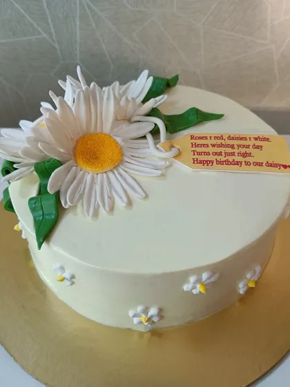 A cheerful daisy-themed cake. This simple white cake is adorned with a large, handcrafted sugar daisy and a cute, personalized message tag.