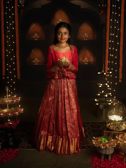 A young girl in a stunning red and gold lehenga holds a diya, her face lit with the joy of Diwali.