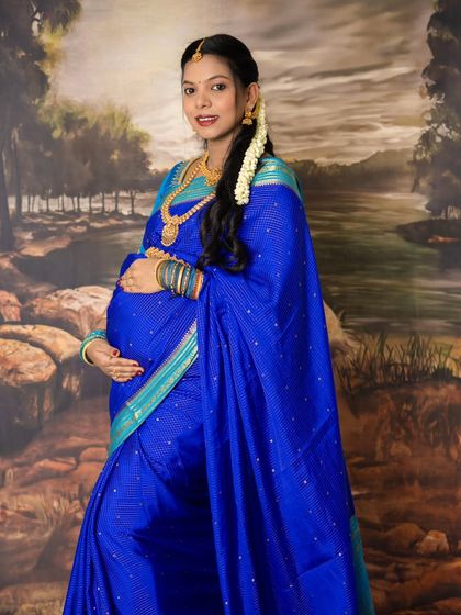 A full-length portrait showcasing the grace of a traditional maternity look. The mom-to-be holds her bump, smiling warmly in her beautiful blue saree.
