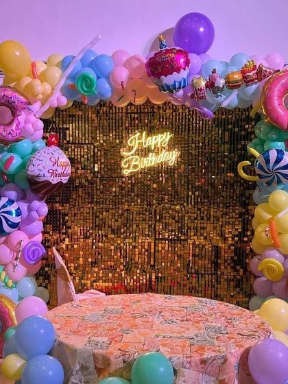 A fun "Candyland" or "Sweet Treats" theme. The decoration features a gold sequin backdrop framed by a colorful balloon arch, which includes large donut, cupcake, and candy foil balloons.