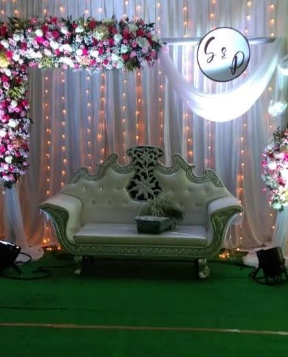 A full wide shot of the engagement stage, showing how the spotlights are used to highlight the floral arrangements and the couple's seating area. The green turf flooring adds a touch of freshness.