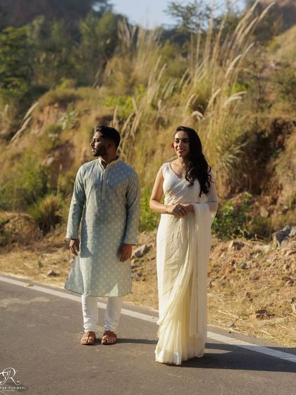 A full-length shot of the couple on a quiet road. Her elegant white saree and his traditional kurta stand out beautifully against the rustic, hilly background of their Delhi engagement shoot.
