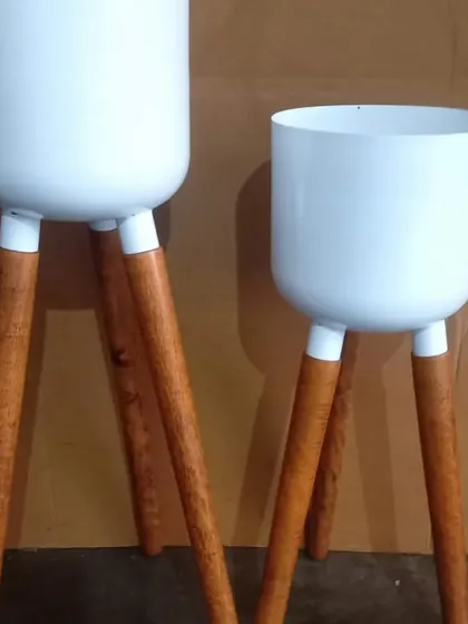 A set of two modern tripod planters. These feature clean white metal pots on sturdy, polished wooden legs, perfect for a Scandinavian or mid-century modern interior style.