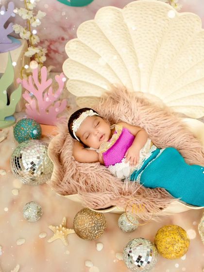 A creative newborn shoot featuring a sleeping baby dressed as a mermaid, nestled inside a large seashell prop with an "under the sea" themed background.