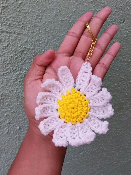 A lovely daisy keychain, held in hand to show its size. The bright yellow center and classic white petals make it a fresh and happy accessory.