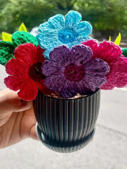 A bouquet of colorful crochet flowers in a pot, held up against a green background. I made this rainbow pot to brighten up any space. Life has enough black and white, so why not add more color?