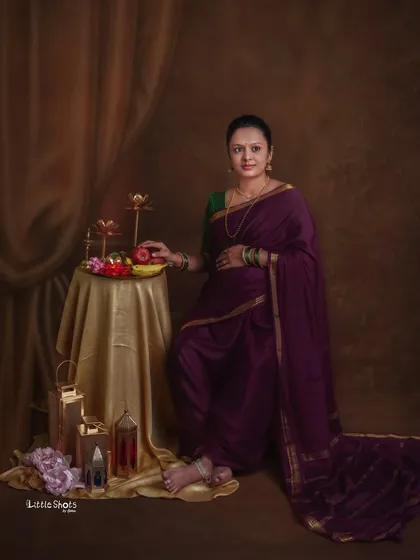 A classic portrait of a mother-to-be in a deep purple saree, styled in a traditional way. She stands next to a table with fruits and brass lamps, evoking a sense of timeless grace.