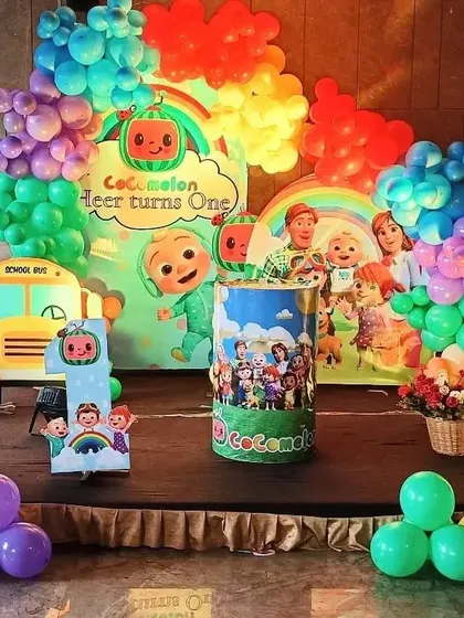 A Cocomelon first birthday party on a stage. This decoration includes a large backdrop, a Cocomelon school bus prop, and a rainbow-colored balloon arch.