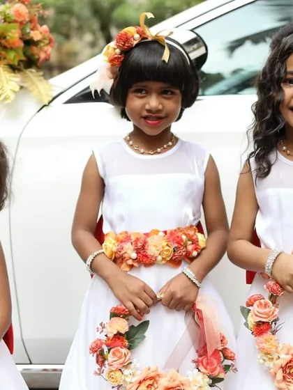 Look at these adorable flower girls. I designed their matching floral accessories, including head wreaths, waist belts, and floral hoops, using a vibrant orange and peach color palette to complement their white dresses and the decorated wedding car. These are the moments that make a wedding unforgettable.