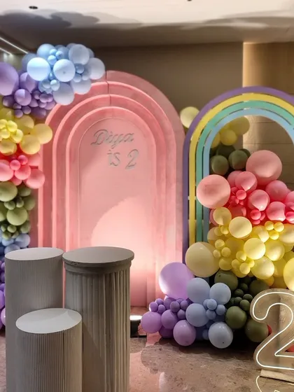 A vibrant rainbow-themed second birthday for Diya. The setup features multiple arches in pastel and rainbow colors, along with a neon number '2', creating a cheerful and multi-dimensional photo backdrop.