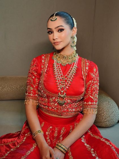 A portrait of bridal serenity. The rich red fabric is complemented by a traditional gold and green choker and a sleek bun adorned with fresh flowers.