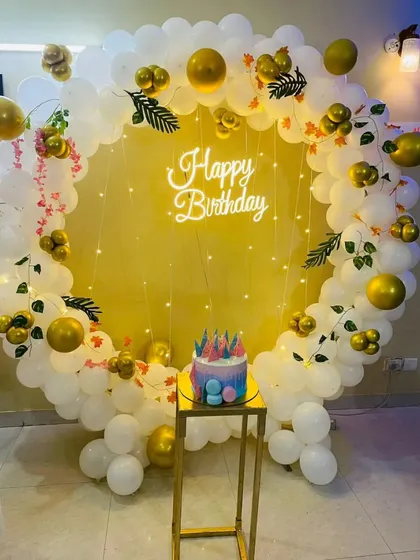 A simple and elegant birthday photo corner with a circular balloon arch in white and gold, a neon sign, and a plinth for the cake.