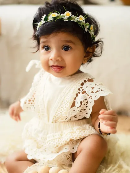 An adorable 8-month-old in a beautiful lace romper and floral headband. The boho studio setup complements her sweet and curious expression perfectly.