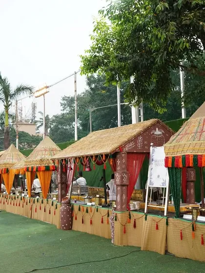A wide-angle shot of our charming village-themed buffet setup for an outdoor wedding. This display features multiple stalls with thatched roofs and traditional decor, perfect for serving authentic Indian cuisine in a rustic and festive atmosphere.