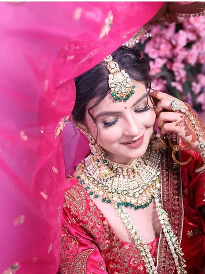 A delicate bridal close-up, with a sheer pink dupatta framing the bride's face. The focus is on her intricate green and gold jewellery and soft makeup.