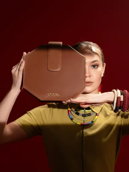 This shot uses the product, an octagonal brown bag, as a prop to create a sense of mystery and style. The model's pose and the strong lighting draw the eye directly to the bag's unique shape and the brand's logo.