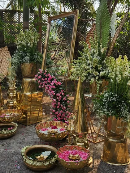 An opulent welcome area with a gold-framed mirror, brass urlis filled with floating flowers, and grand floral arrangements.