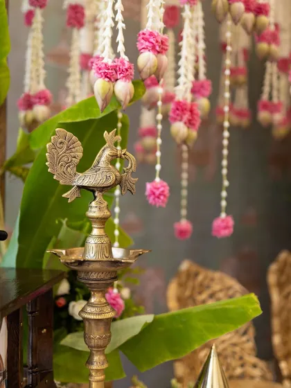 The soft glow of traditional brass lamps, or 'samai', adds a timeless elegance to any ceremony. Here, a classic Annapakshi lamp is set against banana leaves and delicate floral hangings, a small detail that makes a big impact.