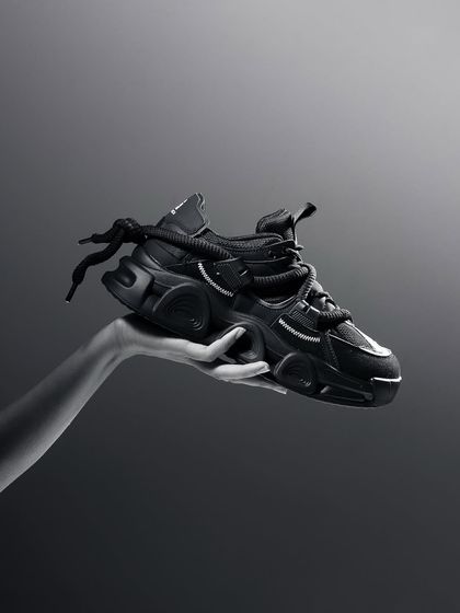 A monochrome shot of a hand holding a futuristic black sneaker. The grayscale tones emphasize the shoe's unique shape and complex design.