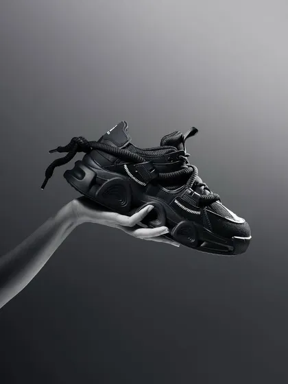 A dramatic black and white shot of a hand holding a chunky black sneaker. The minimalist composition and high-contrast lighting give the product a powerful and iconic look.