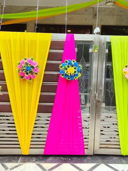 A bright and cheerful gate decoration using yellow, pink, and lime green drapes, each adorned with a colourful floral bouquet.